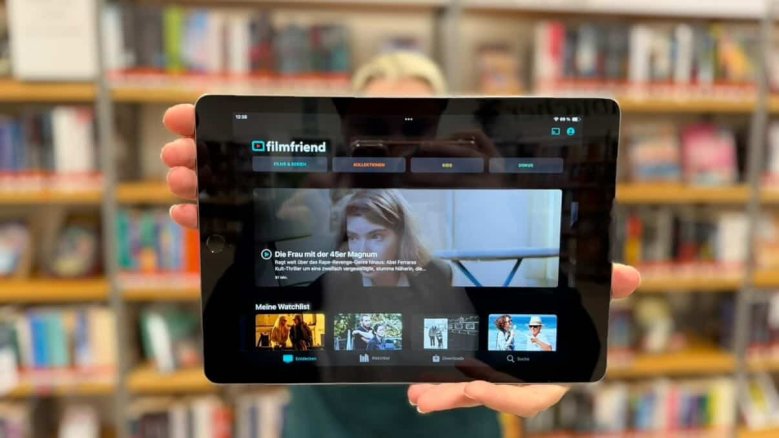 Im Vordergrund ist auf einem Tablet die App von Filmfriend zu sehen. Mit dieser kann man kostenfrei Filme und Serien schauen. Im Hintergrund ist die Bibliothek mit Bücher zu sehen.