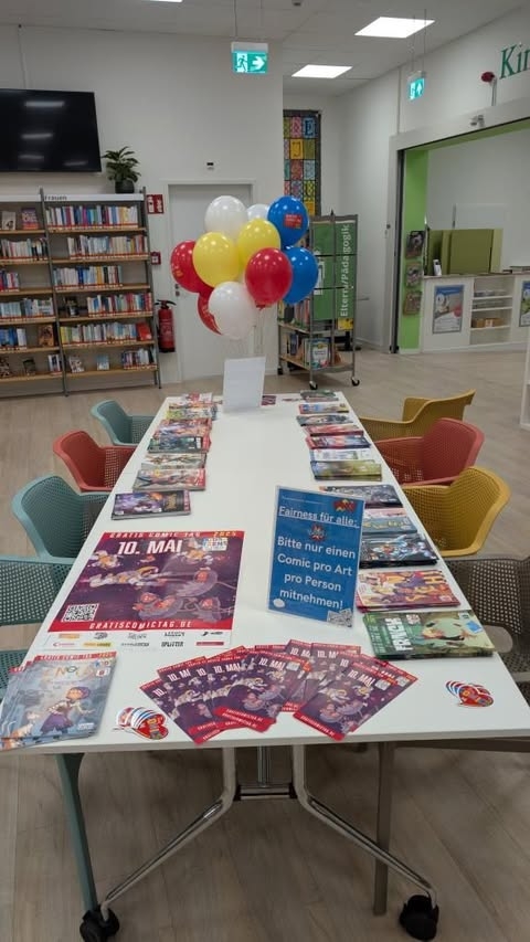 Auf dem Bild ist ein Tisch mit gratis Comics vom Gratiscomictag 2025 zu sehen. Im Hintergrund sind Ballons die verschenkt werden.