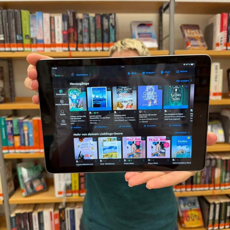 Im Vordergrund ist auf einem Tablet die App von der Onleihe 3.0 zu sehen. Mit dieser kann man E-Books, E-Paper uns sosntiges lesen. Im Hintergrund ist die Bibliothek mit Bücher zu sehen.