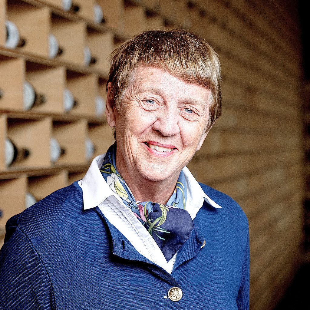 Margarete von Schwarzkopf Margarete von Schwarzkopf in blauem Kostüm, blau-geblumtem Seidenschal, im Hintergrund ein helles Holz-Weinregal mit Weinflaschen