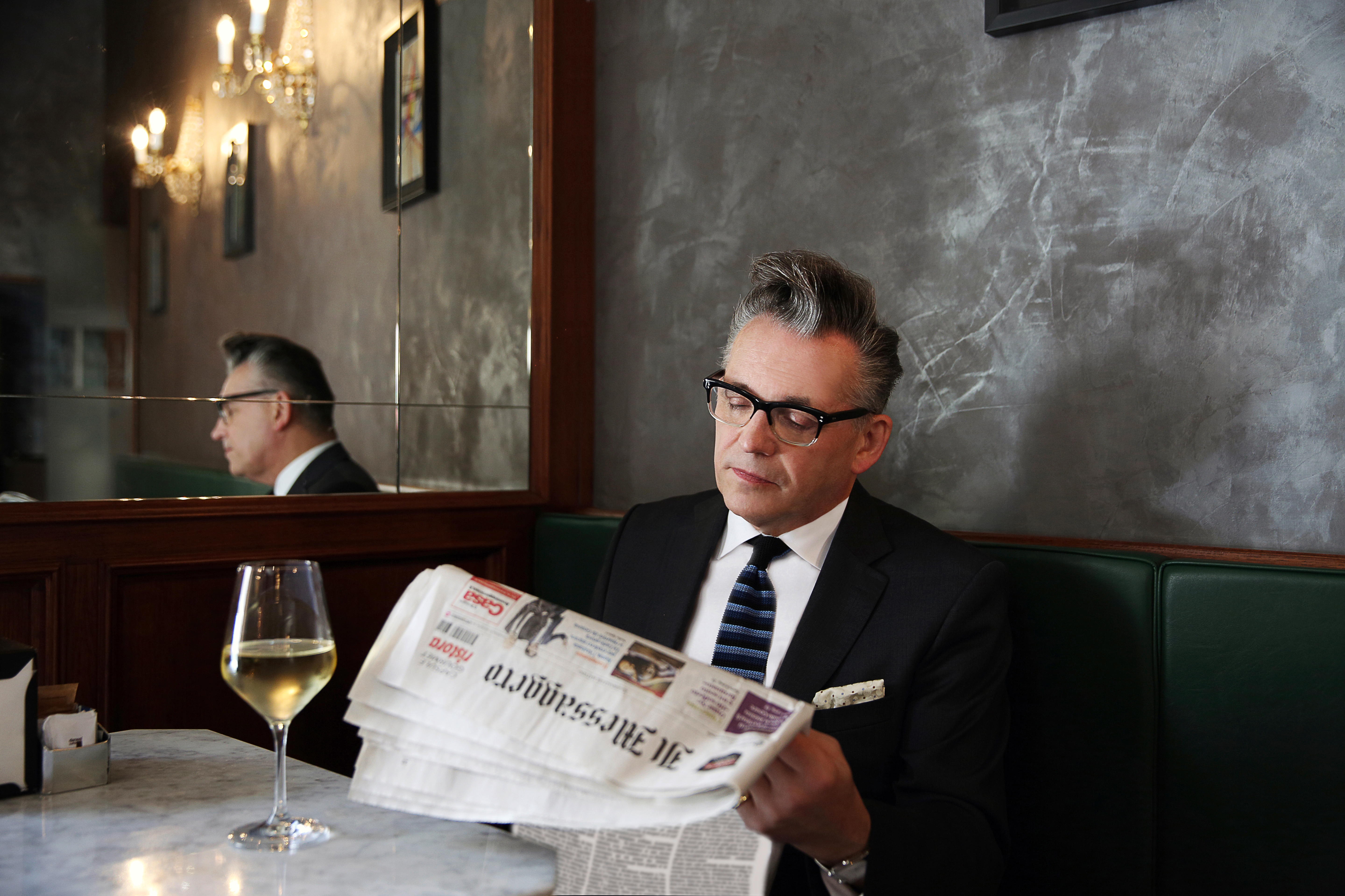 Ein Mann sitzt am Tisch mit einem Glas Wein und einer Zeitung in der Hand.