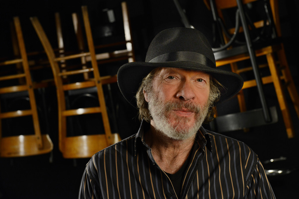 Gerd Köster mit schwarzem Hut, schwarzes Hemd mit Streifen, im Hintergrund hochgestellt Bistrostühle