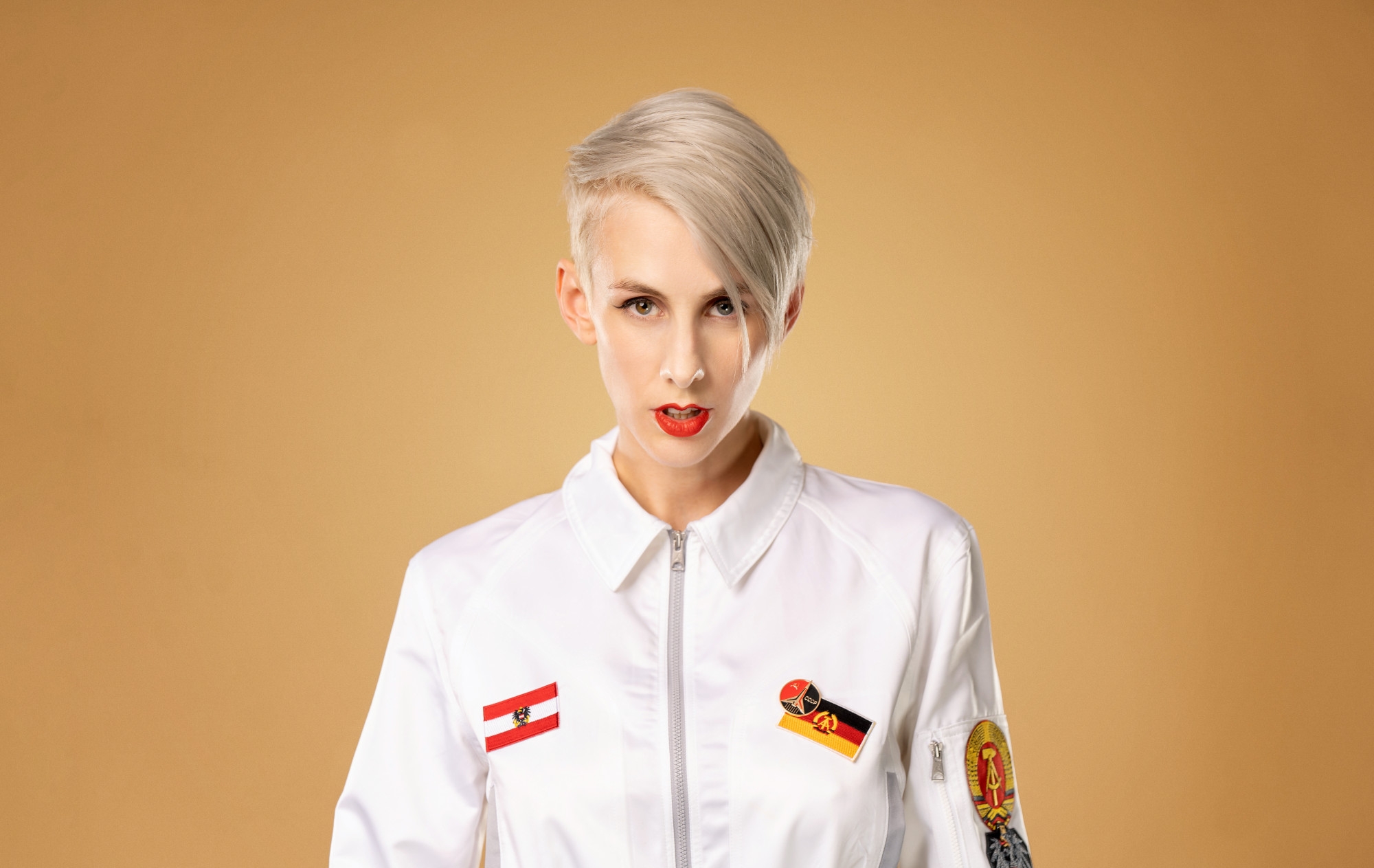 Lisa Eckhart trägt einen weißen Overall und steht vor einem beigen Hintergrund