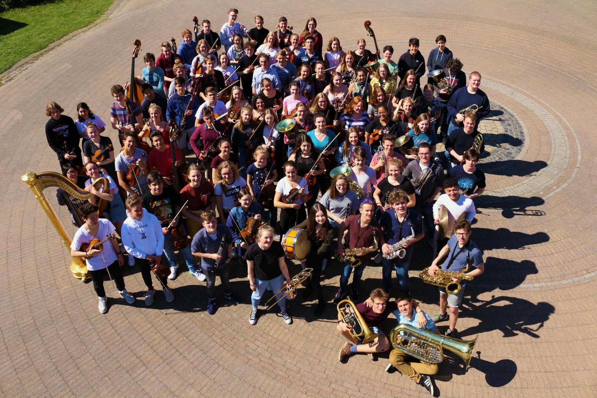Gesamtorchester, bunt gekleidet, mit Instrumenten auf einem Steinboden aus der Luft fotografiert