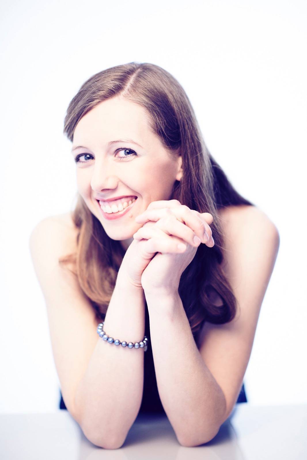 Elvira Bill; braunes langes Har, blaue Augen, lächelt, trägt eine silberne Armkette aus Kugeln, die Ellenbogen auf eine weiße Platte gestützt