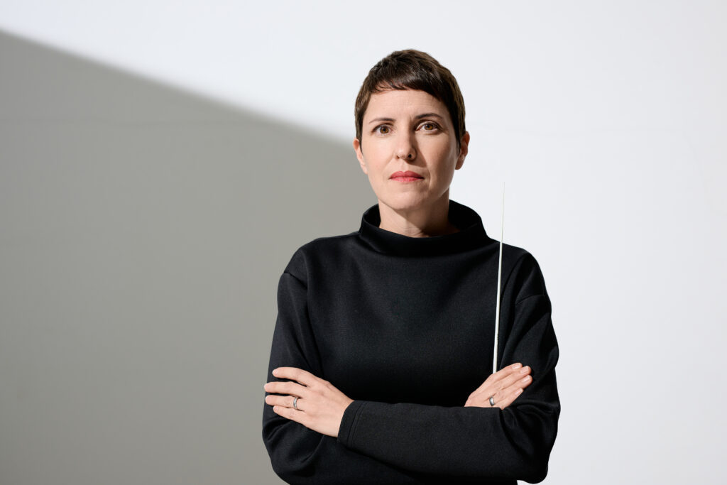 Susanne Blumenthal, braune Kurzhaarfrisur, braune Augen; steht mit verschränkten Armen in einem schwarzen Pullover mit weißen Taktstock in der Hand vor weißem Hintergrund