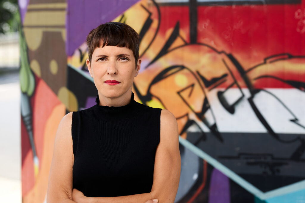 Susanne Blumenthal, braune Kurzhaarfrisur, braune Augen; steht mit verschränkten Armen in einem schwarzen schulterfreien Oberteil mit Stehkragen vor einer Wand im Freien mit bunten Graffitis.