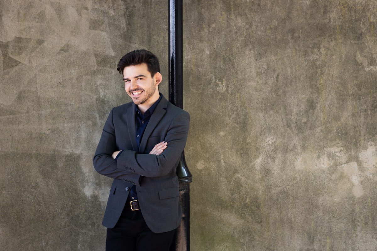 Harry Ogg im antrazitfarbenem Sakko auf einem dunkelblauen Hemd, lehnt an einem schwarzen Metallrohr vor einer beige-braun-grauen Stein-Fassade im Freien