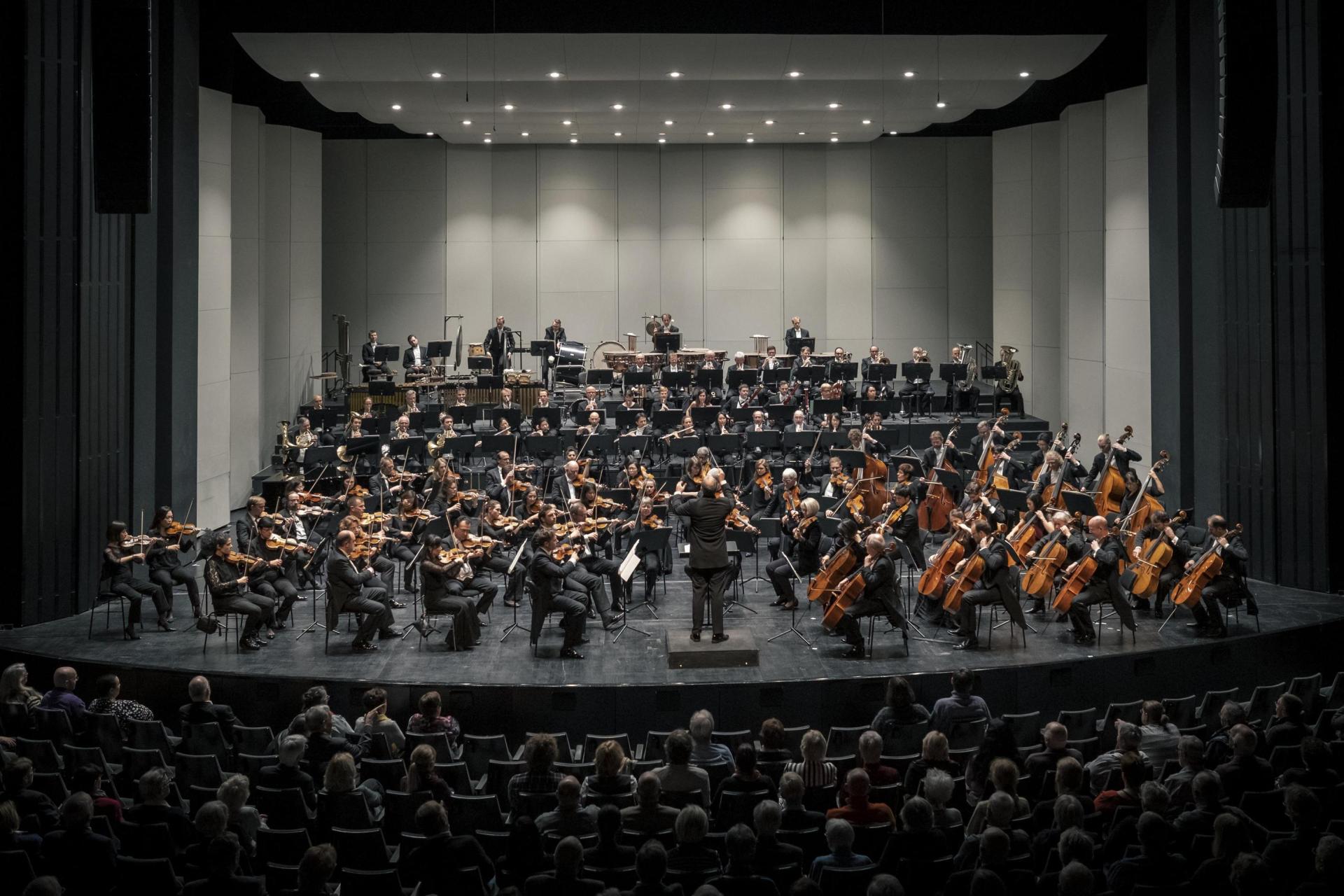 Orchester auf der großen Bühne des Musiktheaters Gelsenkirchen platziert, alle ca. 80 Personen schwarz gekleidet, der Dirigent mittig vor Kopf - dem Orchester zugewandt, vorne die Streicher, mittig die Bläser und hinten das Schlagwerk