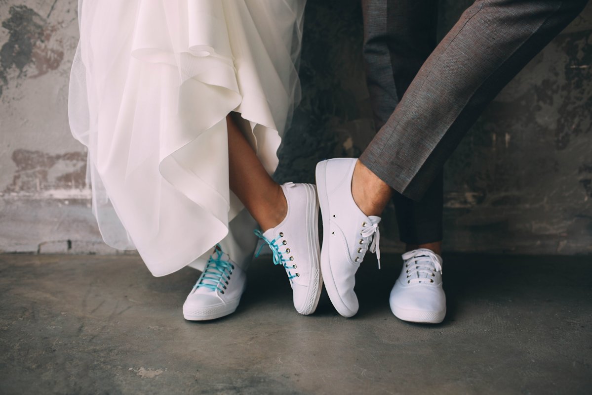 cropped male and female legs wearing white sneakers. Wedding in sneakers Zwei Personen tragen weiße Sneaker – links unter einem fließenden Brautkleid, rechts zu einer grauen Anzughose – vor einer Betonwand.
