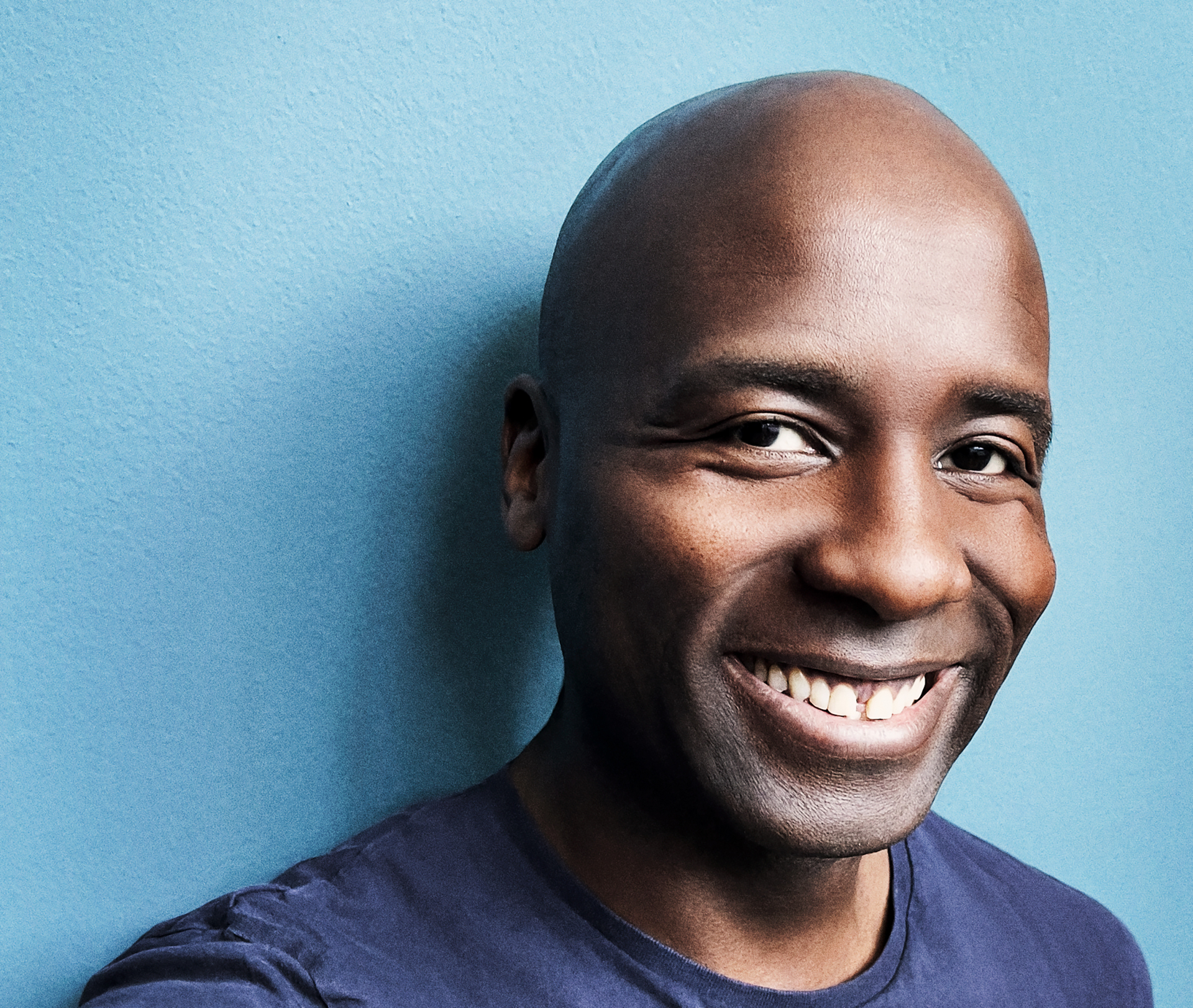 Dave Davis lächelt breit in dunkelblauem Shirt vor einem mittelblauen Hintergrund; Portrait-Foto