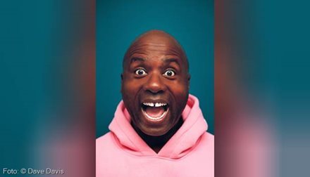 Dave Davis in rosafarbenem Hoodie und freudig überraschtem Gesichtsausdruck vor petrol-farbenem Hintergrund