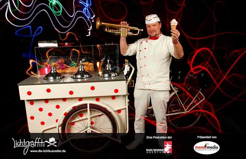 Der Eismann Scotty mit Eis und Trompete in den Händen vor seinem Eiswagen.