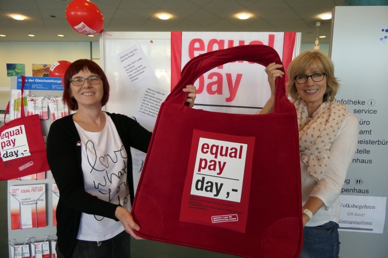 Die rote Tasche steht als Symbol für die Forderung nach Gleichbezahlung: Die Gleichstellungsbeauftragten Karin Lapke-Fernholz (li.) und Petra Römer-Westarp halten im Rathausfoyer Infomaterial zum Equal Pay Day bereit.