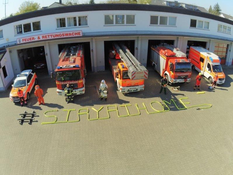 Feuerwehrfahrzeuge und Feuerwehrmitarbeiter vor der Wache. Auf dem Boden steht mit einem Schlauch geschrieben: stay at home