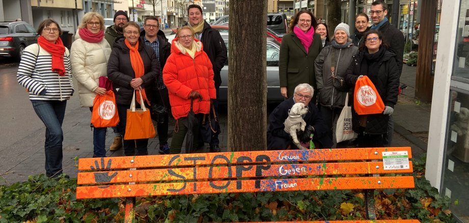 Mehrere Menschen stehen hinter einer orangefarbenen Parkbank