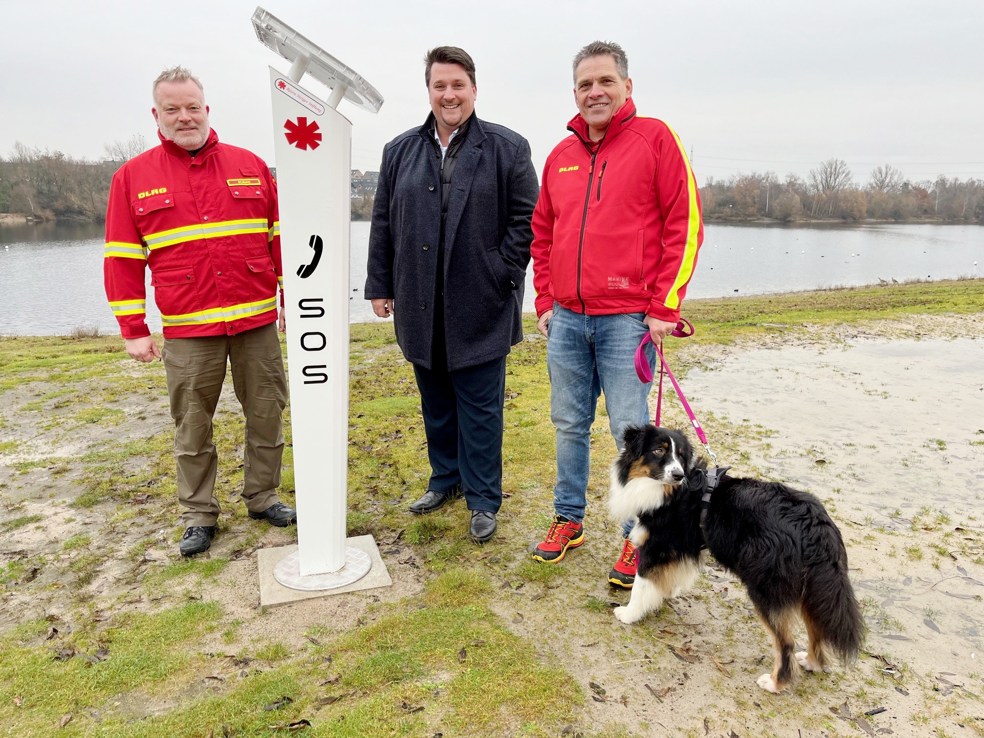 Drei Männer und ein Hund stehen neben einer Notrufsäule