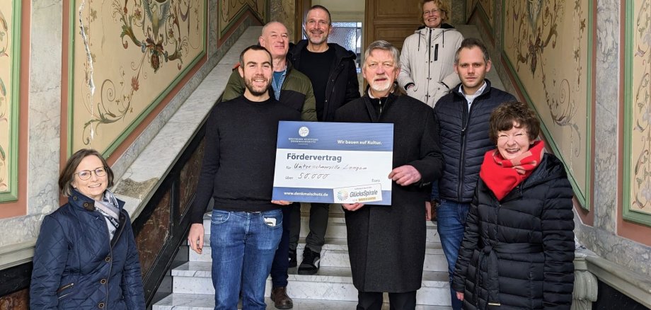 Beate von Berg (Untere Denkmalbehörde Stadt Troisdorf), Sven Riedel, Werner Braun, Andreas Dederichs (Eigentümer), Michale Wangerow (Deutsche Stiftung Denkmalschutz), Marion Grotthof (Eigentümerin), Dennis Teigelkamp (WestLotto), Angelika Müller (Deutsche Stiftung Denkmalschutz) bei der Scheckübergabe im Treppenhaus der Villa Langen.