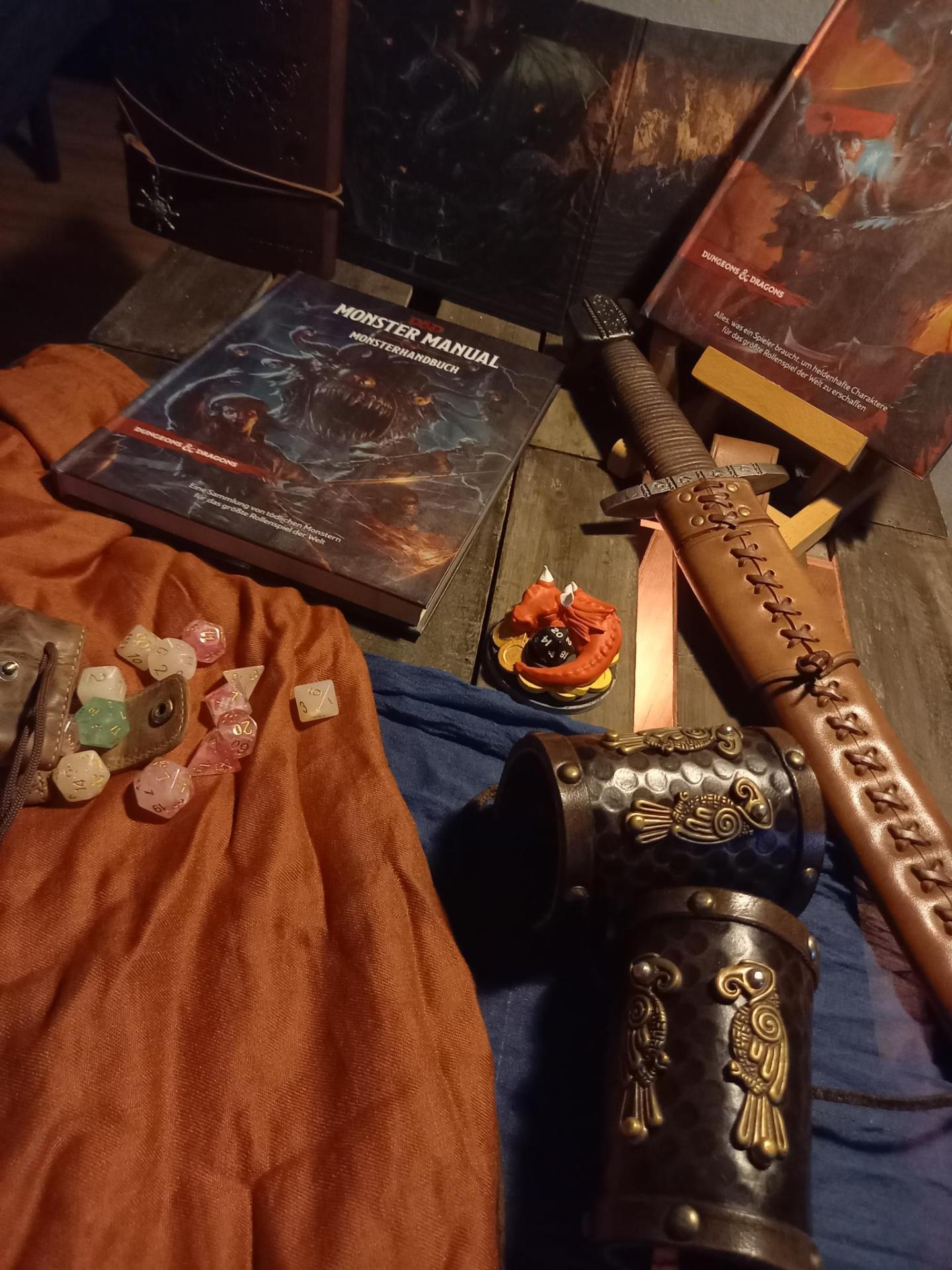 Das Bild zeigt Dungeons & Dragons-Spielmaterial: Bücher, Würfel, ein Schwert, Armschienen und eine Drachenfigur.