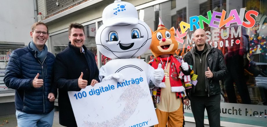 Gemeinsam wird der 100. digitale Antrag gefeiert v.l Fabian Wagner, Leiter der Stabsstelle Digitalisierung, Bürgermeister Alexander Biber, Maskottchen Smarta, die Maus des Karnevalsmuseums und Sebastian Glees, Projektleiter Digitales Rathaus der Stadt Troisdorf.