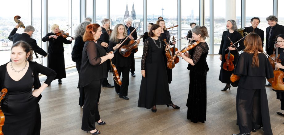 Die Musiker*innen stehen in einem Raum zusdammen. Im Hintergrund sieht man durch die Fensterfront den Kölner Dom