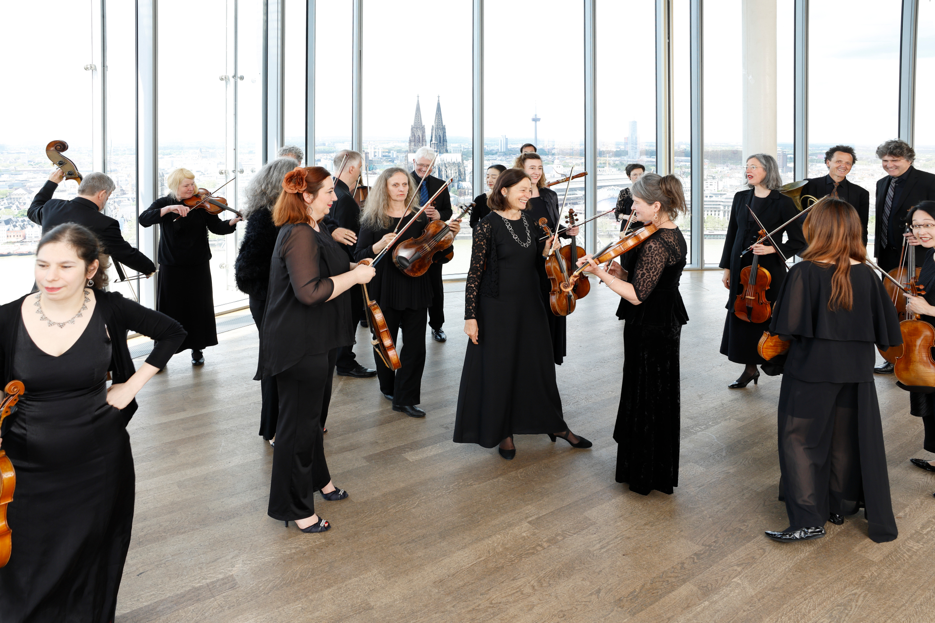Die Musiker*innen stehen in einem Raum zusdammen. Im Hintergrund sieht man durch die Fensterfront den Kölner Dom
