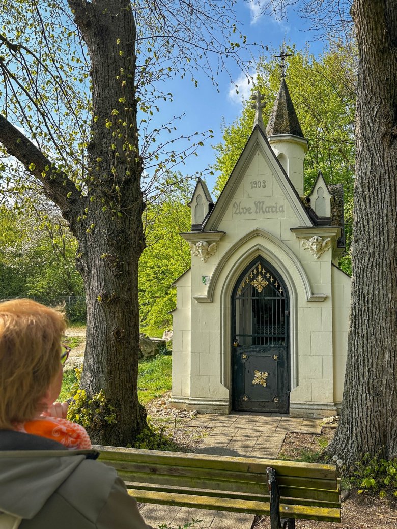 Kleine historische Marienkapelle („Ave Maria“, Baujahr 1903) zwischen Bäumen; im Vordergrund sitzt eine Person auf einer Bank und blickt auf das Gebäude.
