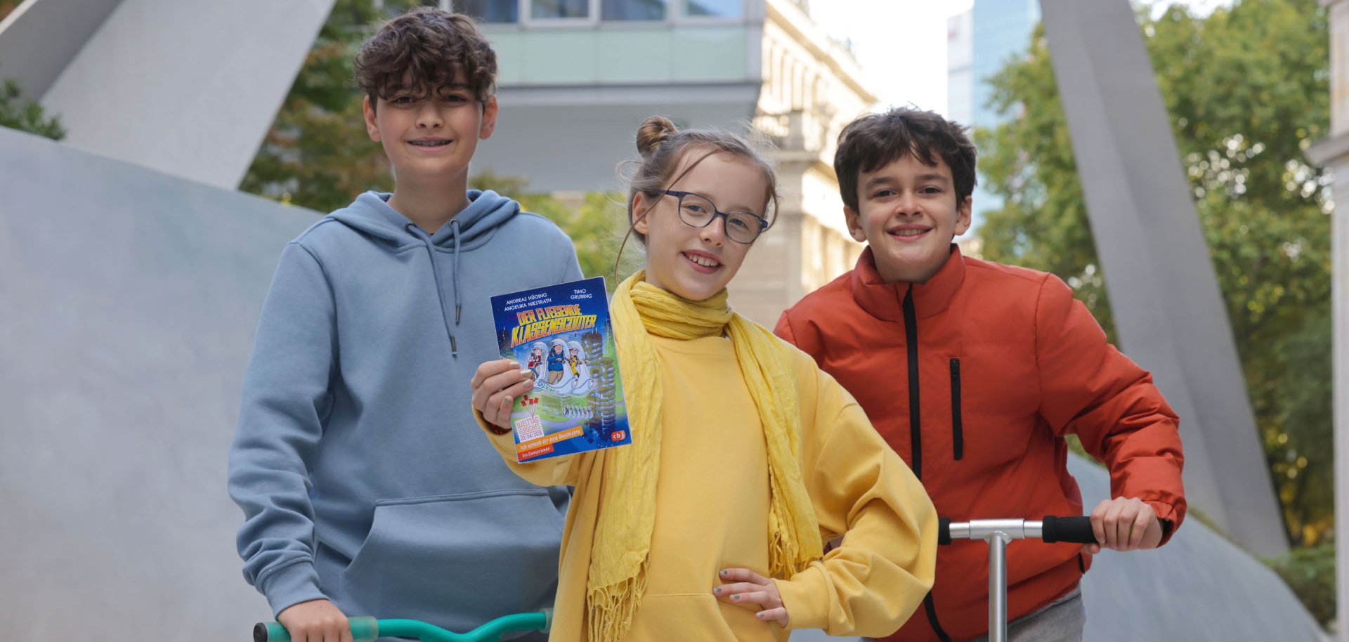 Drei Kinder. Das Mädchen hält ein Buch in die Kamera