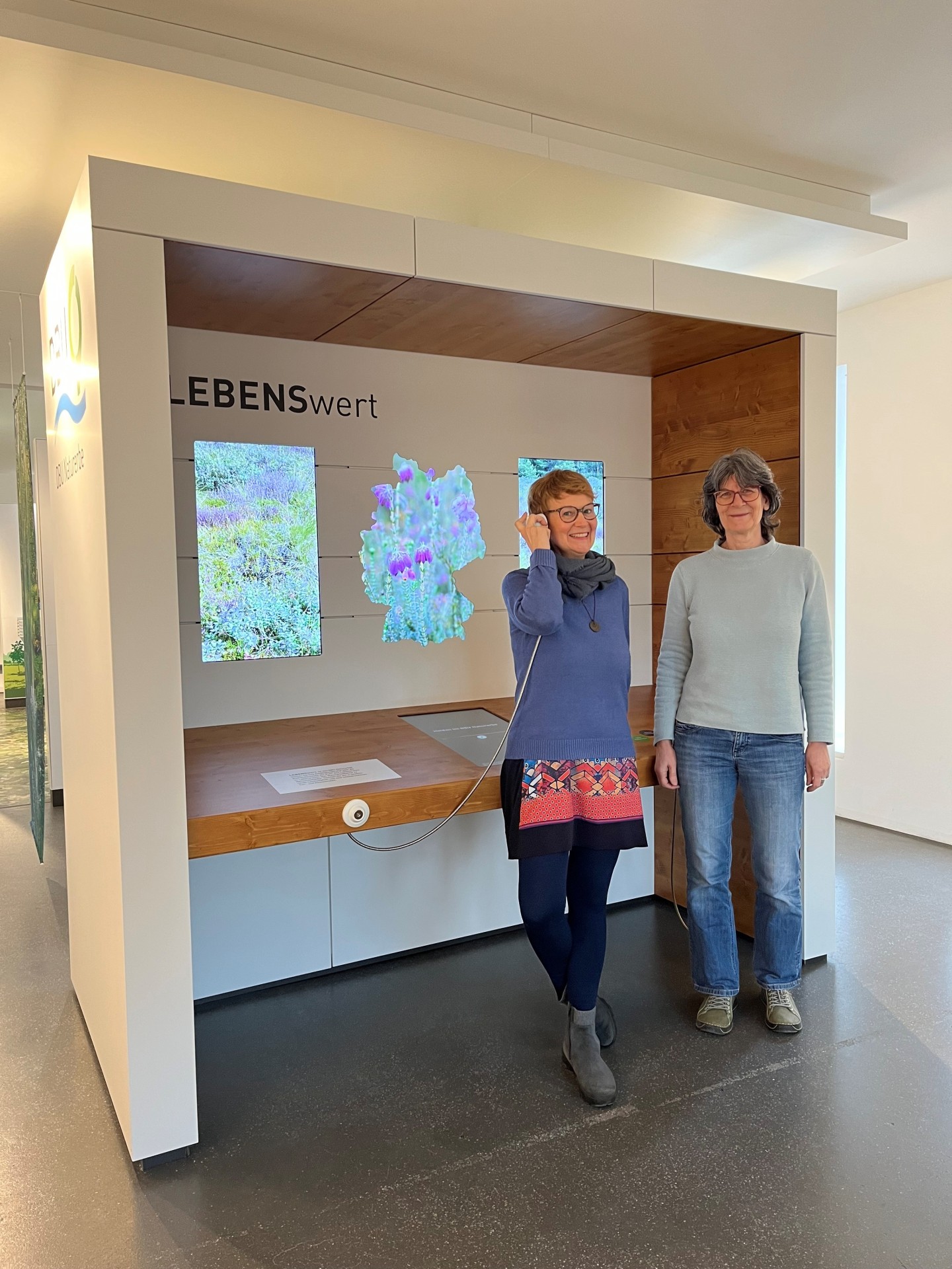 Zwei Frauen stehen vor einer Multivisionswand zum DBU Naturerbe; eine hält lächelnd einen Hörer am Ohr, im Hintergrund sind Naturbilder zu sehen.