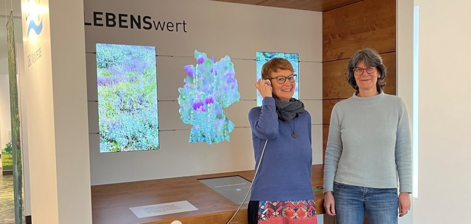 Zwei Frauen stehen vor einer Multivisionswand zum DBU Naturerbe; eine hält lächelnd einen Hörer am Ohr, im Hintergrund sind Naturbilder zu sehen.
