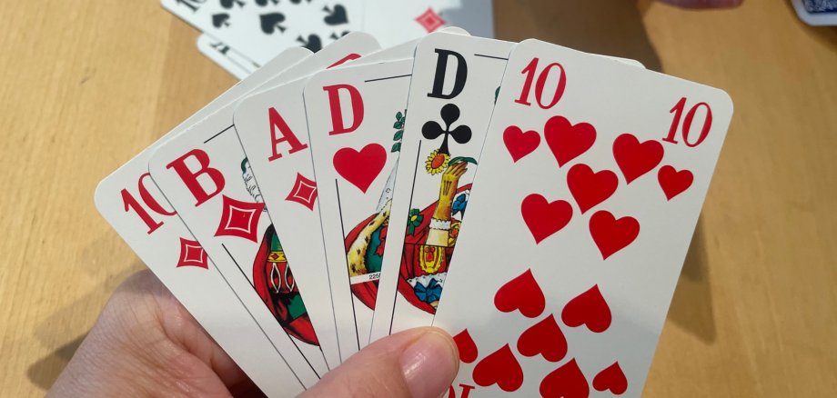 Eine Hand hält mehrere aufgefächerte Spielkarten (u. a. Herz 10 und Bildkarten) über einem Holztisch; im Hintergrund hält eine weitere Person Karten.