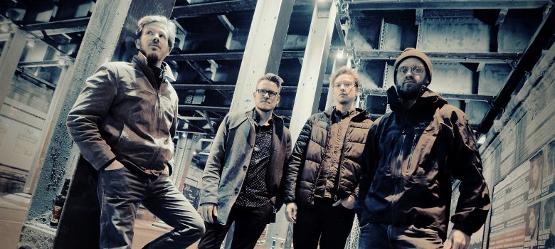 Vier Männer in Graffiti-Halle, dunkle Jacken, einer in hellbraun, Band: Schmid's Huhn.