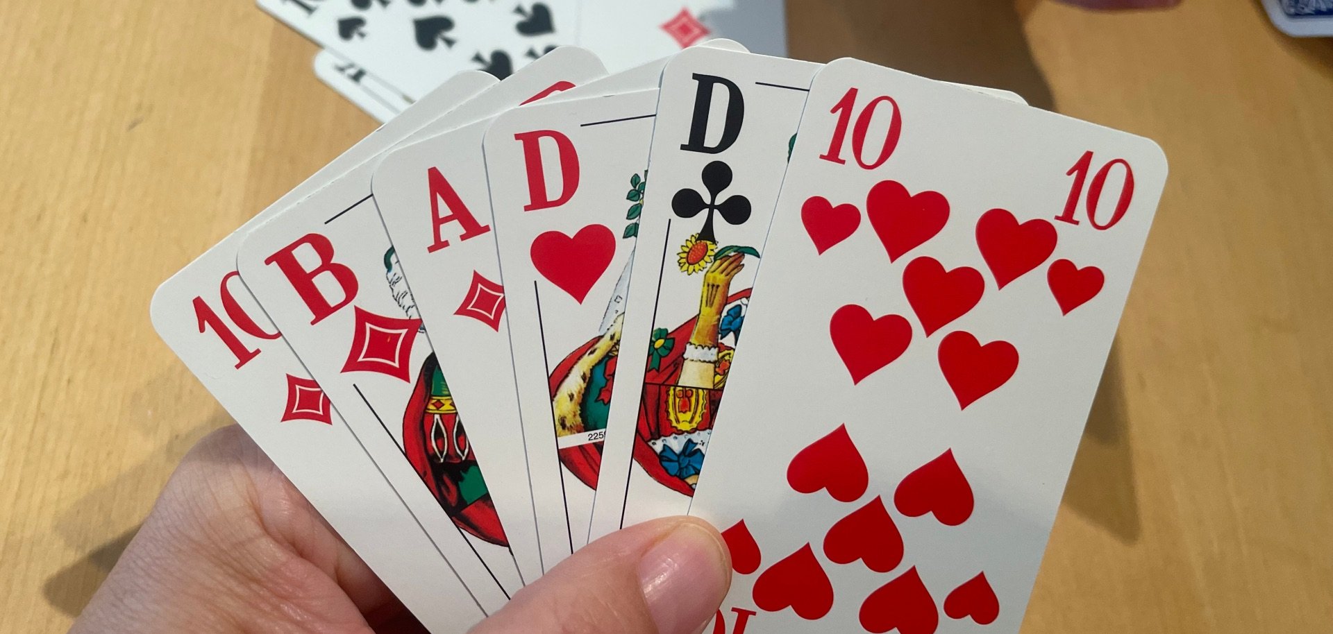 Eine Person hält ein Blatt Doppelkopfkarten in der Hand, während auf dem Tisch bereits ausgespielte Karten liegen und eine weitere Person ihre Karten hält.
