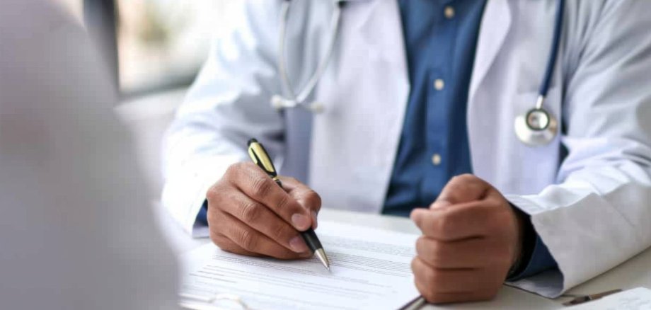 Arzt im Gespräch, hat Stift in der Hand und ein Dokument vor sich auf dem Tisch. 